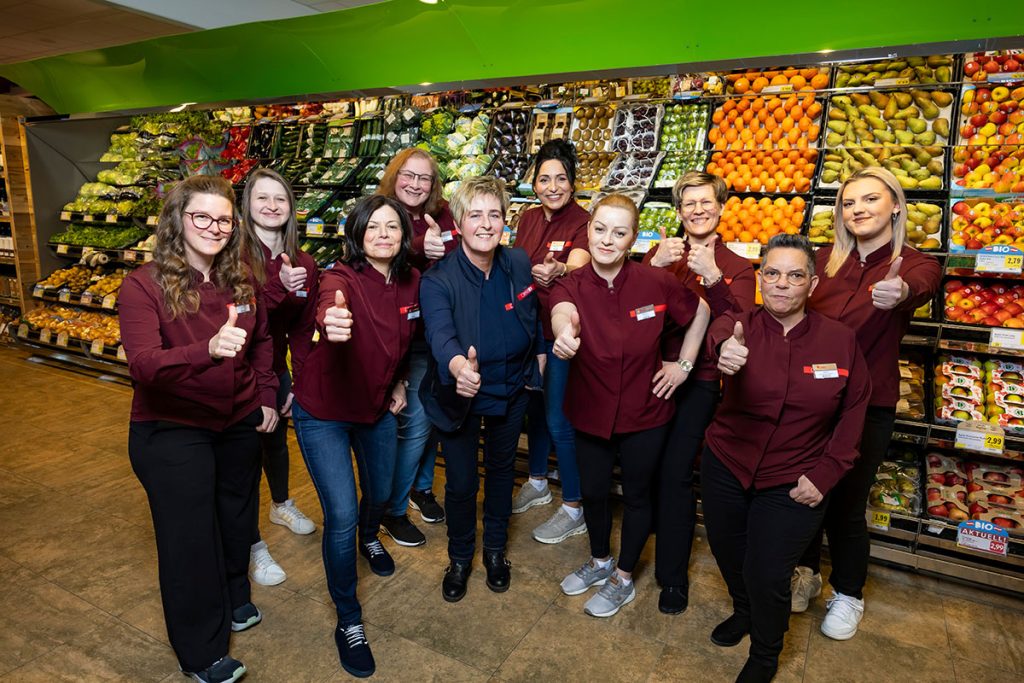 Das Team des SPAR-Marktes Rottenmann steht gemeinsam im Obst- und Gemüsebereich vor gefüllten Regalen mit frischem Obst und Gemüse. Die Mitarbeiterinnen und Mitarbeiter tragen rote SPAR-Uniformen und zeigen lächelnd den Daumen nach oben. In der Mitte steht die Marktleiterin des SPAR Rottenmann.