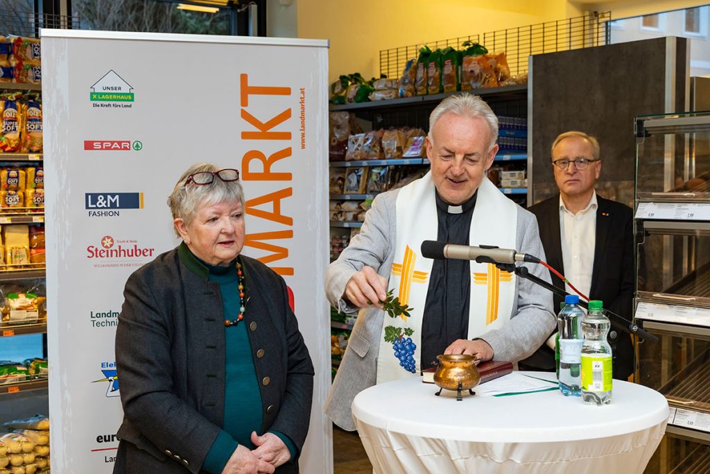 Ein Geistlicher segnet während der Eröffnungsfeier den SPAR-Markt Rottenmann. Er steht an einem Stehtisch mit Mikrofon und Weihwassergefäß und führt die Segnung durch. Daneben stehen weitere Gäste, im Hintergrund sind Verkaufsregale sowie ein Banner der Landmarkt KG zu sehen.