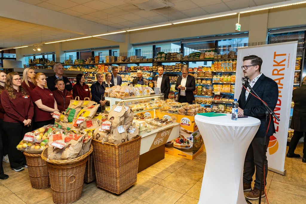 Der Bürgermeister von Rottenmann hält während der Eröffnungsfeier eine Ansprache im SPAR-Markt Rottenmann. Vor ihm stehen Mitarbeiterinnen des Marktes und Gäste der Veranstaltung, im Vordergrund sind Verkaufsstände mit Obst und regionalen Produkten zu sehen. Im Hintergrund befinden sich Regale mit verschiedenen Lebensmitteln.