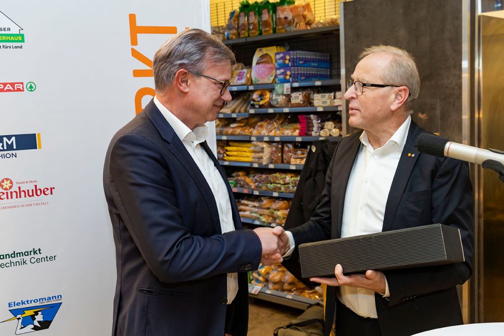 Der Geschäftsführer der Landmarkt KG (rechts) überreicht während der Eröffnungsfeier im SPAR-Markt Rottenmann ein Geschenk an einen SPAR-Vertreter. Die beiden Männer reichen sich die Hand. Im Hintergrund sind Verkaufsregale mit Lebensmitteln sowie ein Banner der Landmarkt KG zu sehen.