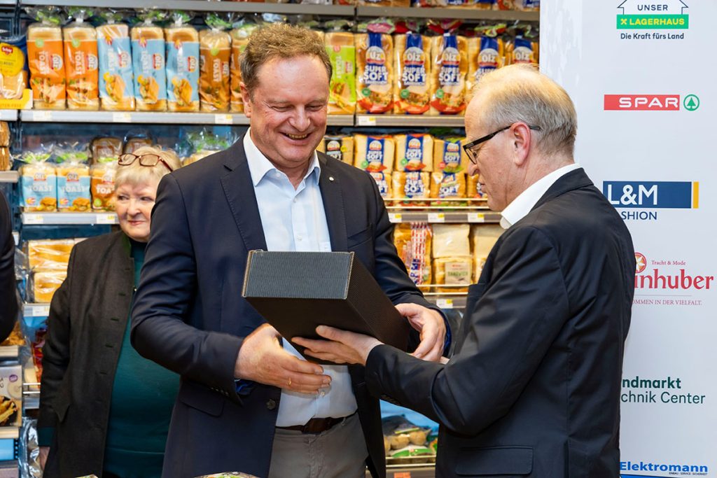 Der Geschäftsführer der Landmarkt KG überreicht während der Eröffnungsfeier im SPAR-Markt Rottenmann ein Geschenk an einen Vertreter von SPAR. Im Hintergrund sind Regale mit Brot und Backwaren sowie ein Banner mit Logos der Landmarkt-Unternehmensbereiche zu sehen.