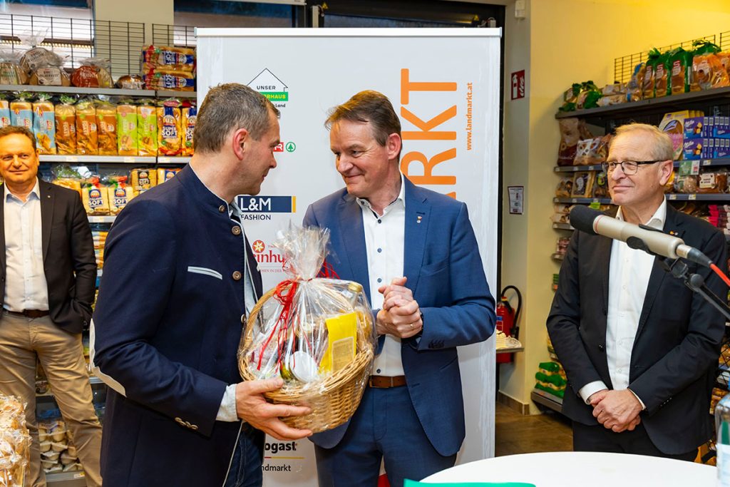 Der Bereichsleiter Landmarkt SPAR (im blauen Anzug) überreicht während der Eröffnungsfeier im SPAR-Markt Rottenmann dem ehemaligen Inhaber des Marktes einen Geschenkkorb. Daneben steht der Geschäftsführer der Landmarkt KG am Mikrofon. Im Hintergrund sind Verkaufsregale mit Lebensmitteln sowie ein Banner der Landmarkt KG zu sehen.