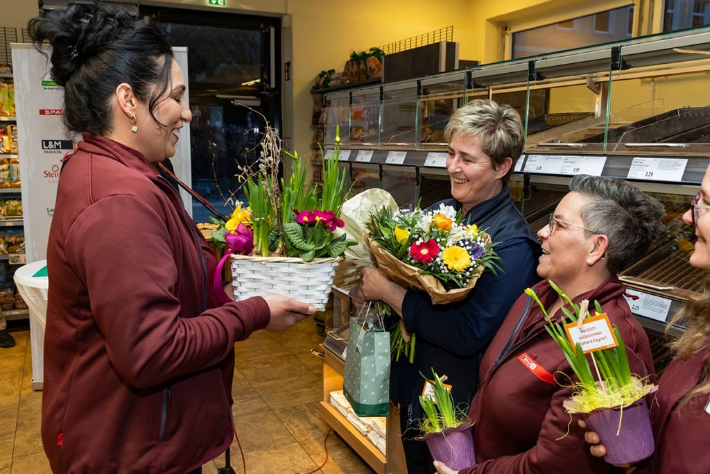 Eine Mitarbeiterin des SPAR-Marktes überreicht einer Kollegin einen dekorativen Frühlingskorb mit Blumen. Die Beschenkte hält einen Blumenstrauß in der Hand und wird von weiteren Mitarbeiterinnen im SPAR-Team begrüßt. Die Szene findet im Markt vor der Backwarenabteilung statt.