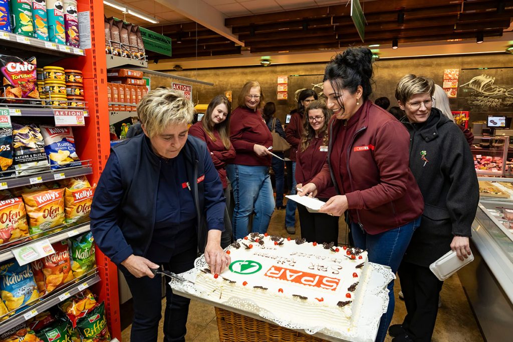 Mitarbeiterinnen des SPAR-Marktes Rottenmann schneiden gemeinsam eine große SPAR-Torte an. Rundherum stehen Kolleginnen aus dem Team und beobachten den Moment. Die Aufnahme entstand im Markt zwischen den Verkaufsregalen während der Eröffnungsfeier.