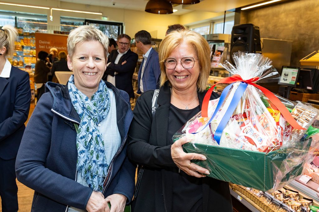 Zwei Marktleiterinnen aus anderen Landmarkt-SPAR-Filialen stehen im Marktbereich eines SPAR-Supermarktes und lächeln in die Kamera. Eine der beiden hält einen Geschenkkorb mit verschiedenen Lebensmitteln und einer Schleife in den Farben Rot, Weiß und Blau. Im Hintergrund sind weitere Gäste sowie Verkaufsbereiche des Marktes zu sehen.