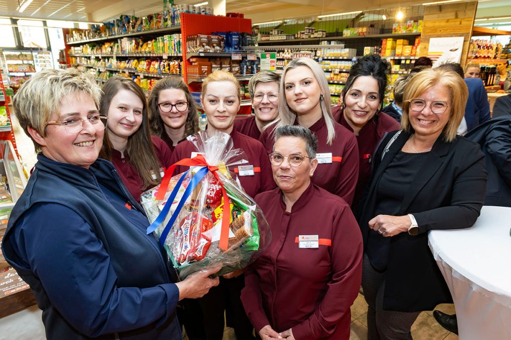 Mehrere Mitarbeiterinnen des SPAR-Marktes Rottenmann stehen gemeinsam im Verkaufsbereich vor gefüllten Regalen mit Lebensmitteln. Eine Person überreicht der Marktleiterin einen Geschenkkorb mit verschiedenen Produkten und einer Schleife. Die Mitarbeiterinnen tragen rote SPAR-Uniformen und lächeln in die Kamera.