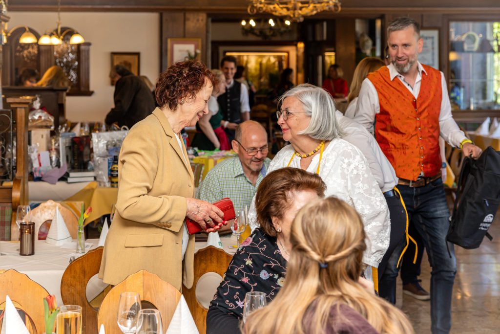 Mehrere Gäste unterhalten sich in einem festlich dekorierten Restaurant bei einer Veranstaltung. Zwei ältere Frauen stehen lächelnd im Gespräch, während andere Besucher an den Tischen sitzen. Im Hintergrund bewegt sich ein Mann in orangefarbener Weste durch den Saal, der mit warmem Licht und Holzelementen gemütlich eingerichtet ist.