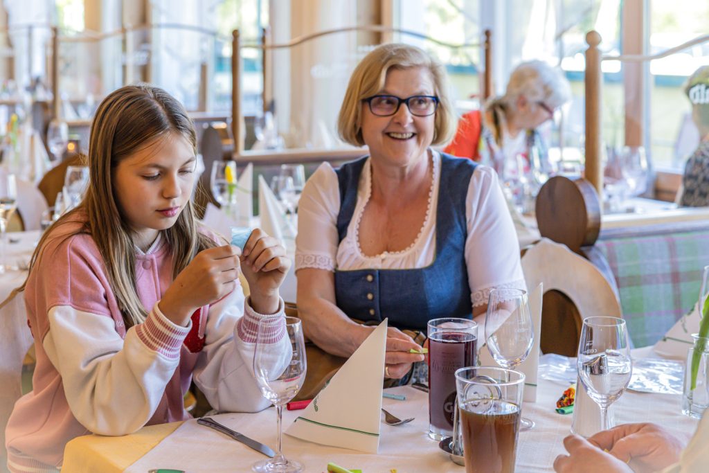 Ein Mädchen in rosa-weißem Pullover sitzt gemeinsam mit einer lächelnden Frau in Tracht an einem gedeckten Tisch in einem Restaurant. Während das Mädchen etwas in den Händen betrachtet, schaut die Frau freundlich zur Seite. Auf dem Tisch stehen Gläser, Servietten und Getränke, im Hintergrund sind weitere Gäste zu sehen.