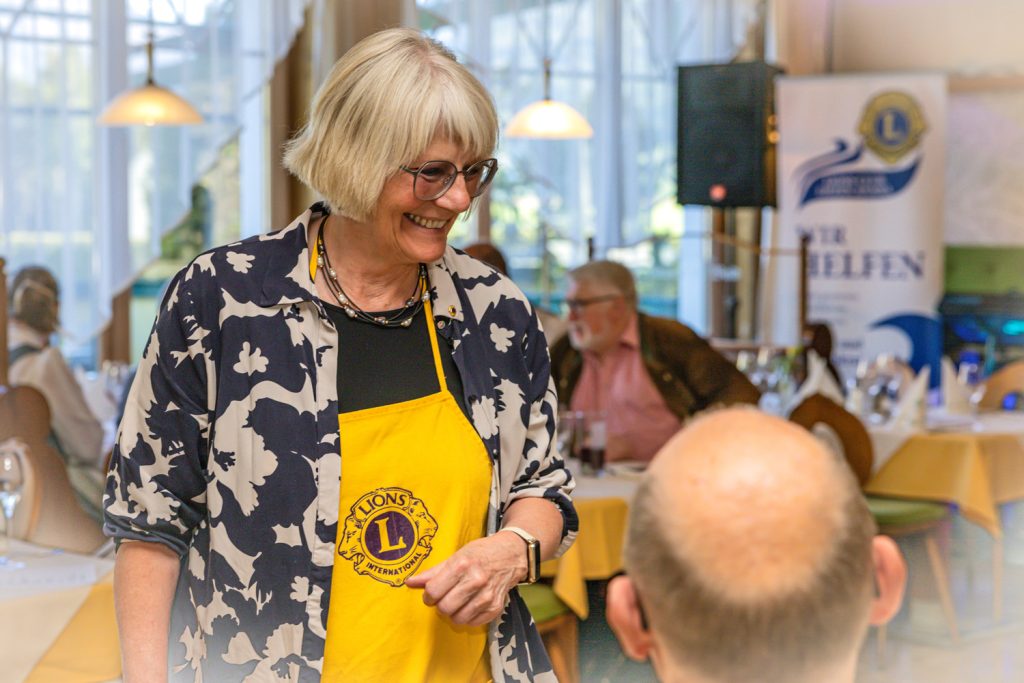 Eine lächelnde Frau mit heller Kurzhaarfrisur und Brille trägt eine gelbe Schürze mit dem Logo von Lions International und unterhält sich bei einer Veranstaltung mit einer sitzenden Person. Im Hintergrund sind gedeckte Tische und ein Banner des Lions Club Liezen Anasia zu sehen.