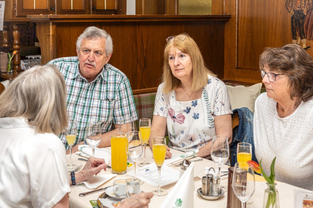 Vier Personen sitzen gemeinsam an einem gedeckten Tisch in einem Restaurant und führen ein aufmerksam wirkendes Gespräch. Auf dem Tisch stehen Gläser mit Orangensaft, Geschirr und eine kleine Blumenvase. Die Atmosphäre wirkt freundlich, ruhig und gesellig.