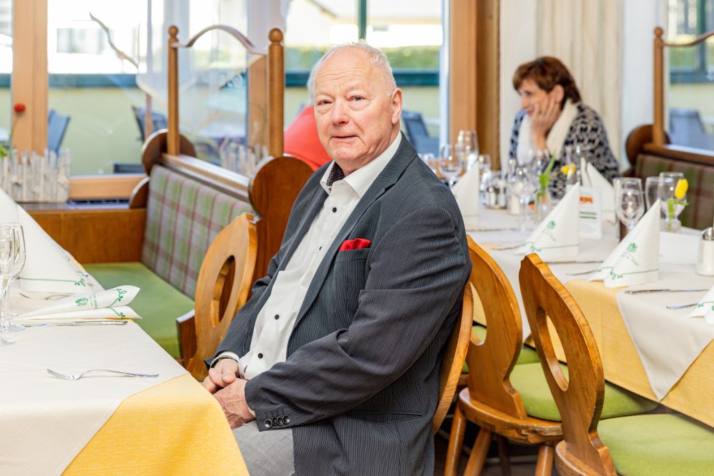 Ein älterer Mann im dunklen Sakko sitzt an einem festlich gedeckten Tisch in einem Restaurant und blickt freundlich in die Kamera. Im Hintergrund sind weitere gedeckte Tische und eine Frau zu sehen. Die Atmosphäre wirkt ruhig und elegant.