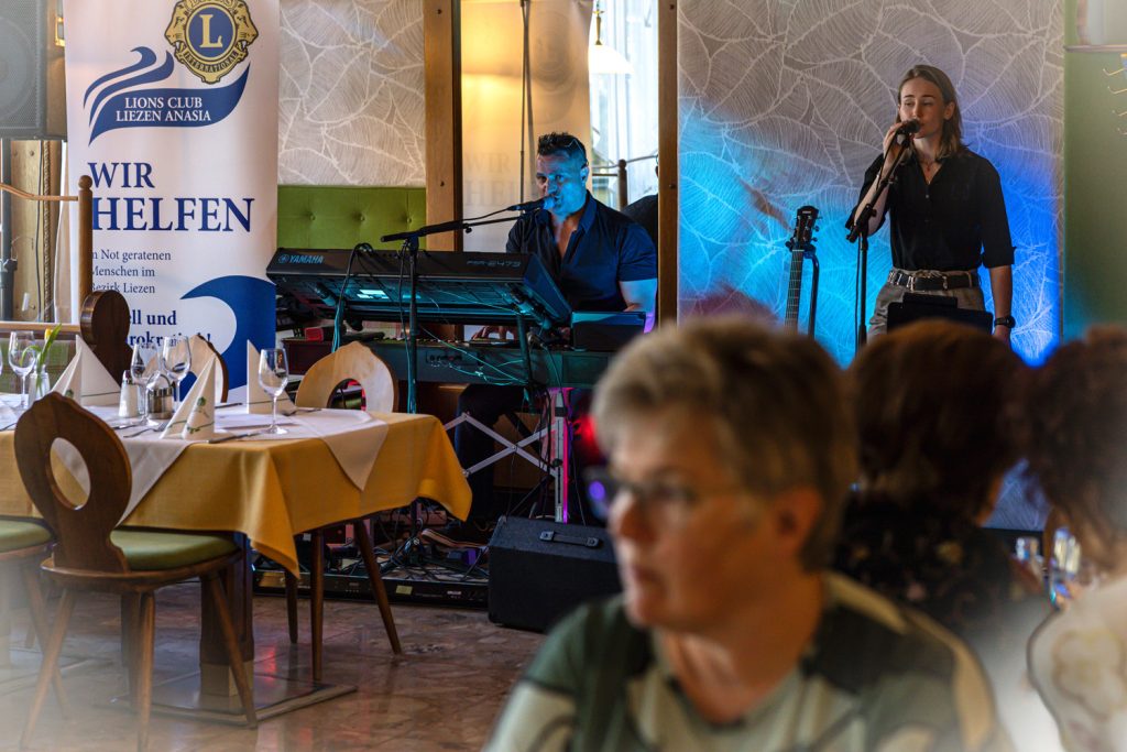 Zwei Musiker treten in einem Restaurant vor Gästen auf. Ein Mann sitzt am Keyboard und singt in ein Mikrofon, während eine junge Frau daneben am Mikrofon steht. Im Vordergrund sitzen Gäste an gedeckten Tischen, und die Bühne ist mit blauem Licht beleuchtet.