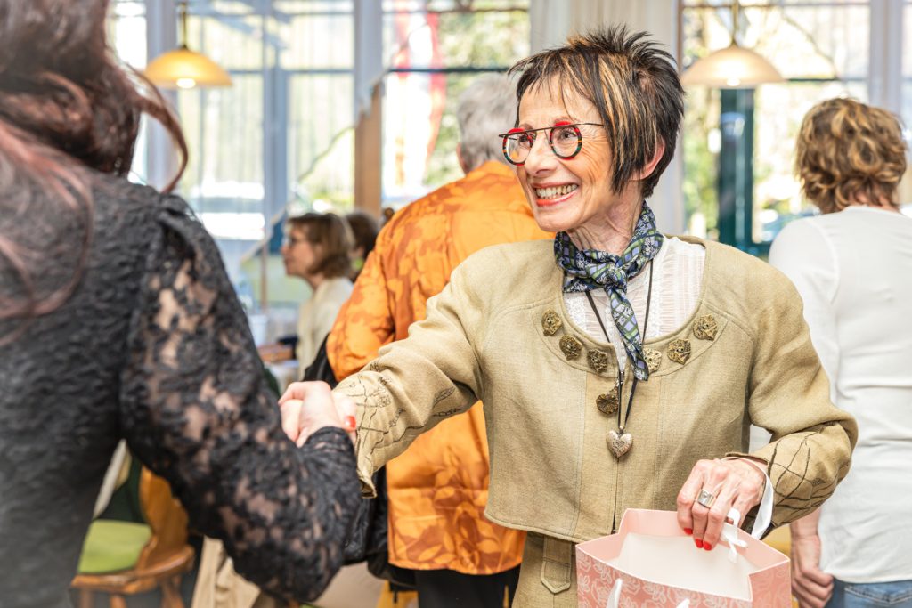 Eine elegant gekleidete ältere Frau mit roter Brille lächelt freundlich und begrüßt eine andere Person mit Handschlag in einem Restaurant. In der einen Hand hält sie eine rosa Geschenktasche. Im Hintergrund sind weitere Gäste und eine helle, gesellige Atmosphäre zu sehen.