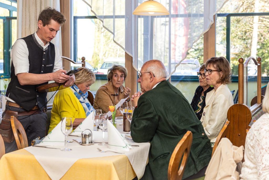 Ein Kellner steht neben einem gedeckten Tisch in einem Restaurant und bedient eine Gruppe von Gästen. Mehrere ältere Personen sitzen zusammen, unterhalten sich und schauen auf Speisekarten oder den Kellner. Die Atmosphäre wirkt ruhig, freundlich und gesellig.