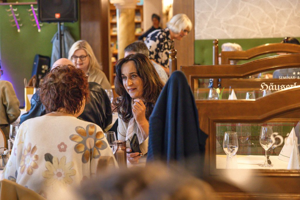 Zwei Frauen sitzen in einem gemütlichen Restaurantbereich an einem Tisch und unterhalten sich aufmerksam miteinander. Eine der Frauen hält ein Smartphone in der Hand und lächelt freundlich, während im Hintergrund weitere Gäste und eine Person mit Geschenktasche zu sehen sind. Die Stimmung wirkt warm, entspannt und gesellig.