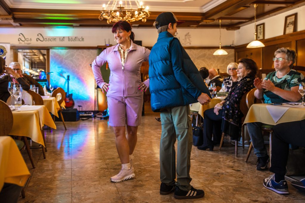 Zwei Personen präsentieren Sportmode auf einem Laufsteg in einem traditionellen Gasthaus. Eine Frau trägt ein sportliches Outfit in Rosa mit Sneakern, daneben steht ein junger Mann in blauer Outdoorjacke, weiter Hose und Cap. Die Gäste sitzen an den Tischen und applaudieren der Modenschau.