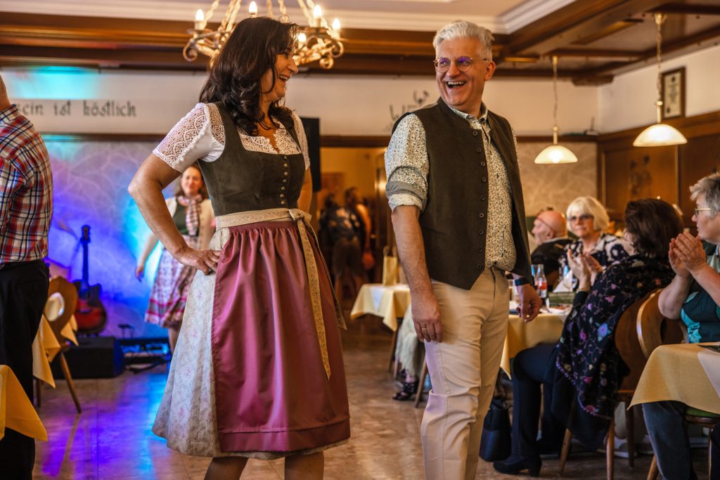 Ein lachendes Paar präsentiert Trachtenmode auf einem Laufsteg in einem traditionellen Gasthaus. Die Frau trägt ein elegantes Dirndl in Rosé- und Olivtönen mit Spitzenbluse, der Mann kombiniert eine gemusterte Trachtenweste mit hellem Hemd und beiger Hose. Die Gäste sitzen an den Tischen und applaudieren der Modenschau.