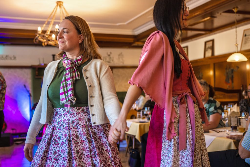Zwei Frauen in floralen Trachtenoutfits gehen Hand in Hand über den Laufsteg einer Modenschau. Eine trägt eine helle Strickjacke mit gemustertem Schal, die andere ein rosafarbenes Dirndl mit passendem Schultertuch. Im Hintergrund sorgen bunte Lichter und die Gäste im Gasthaus für eine festliche Atmosphäre.