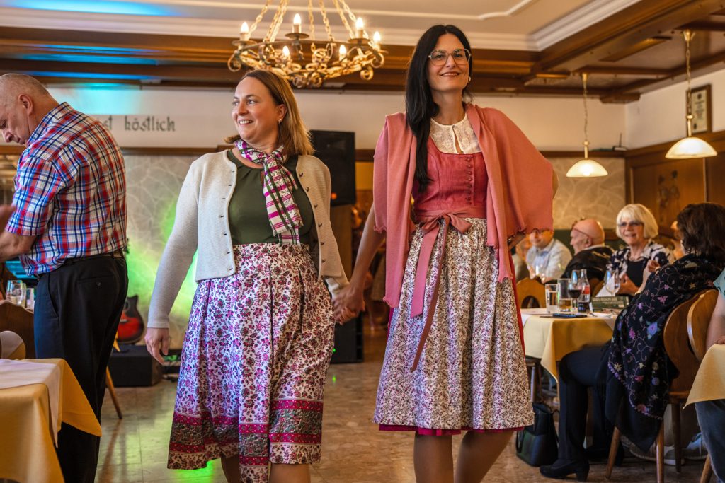 Zwei Frauen präsentieren gemeinsam Trachtenmode auf einem Laufsteg in einem traditionellen Gasthaus. Eine trägt ein florales Kleid mit heller Strickjacke und gemustertem Schal, die andere ein rosafarbenes Dirndl mit passendem Schultertuch. Im Hintergrund sitzen Gäste an gedeckten Tischen und verfolgen die Modenschau.