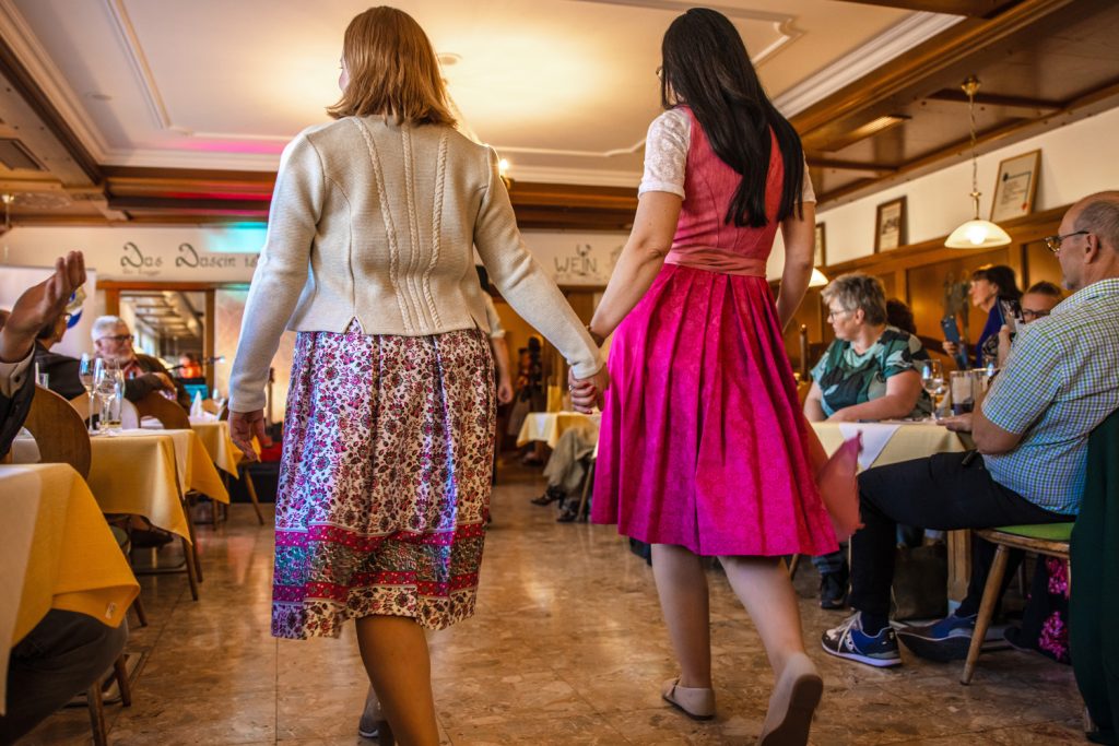 Zwei Frauen gehen Hand in Hand über den Laufsteg einer Modenschau in einem gemütlichen Restaurant. Eine trägt ein florales Trachtenkleid mit heller Strickjacke, die andere ein kräftig pinkes Dirndl. Die Gäste sitzen links und rechts an den Tischen und verfolgen die Präsentation aufmerksam.