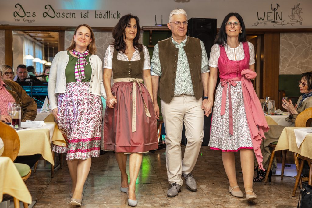 Vier Personen präsentieren gemeinsam elegante Trachtenmode bei einer Modenschau im Gastraum eines Restaurants. Die Frauen tragen verschiedene Dirndl in floralen Mustern und harmonischen Farben, der Mann ein traditionelles Outfit mit Weste. Hand in Hand gehen sie über den Laufsteg, während die Gäste die Präsentation aufmerksam verfolgen.