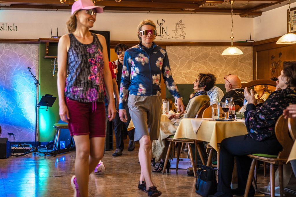 Mehrere Personen präsentieren sportliche Freizeit- und Outdoorbekleidung von Sport Spanner bei einer Modenschau. Zu sehen sind funktionelle Shirts, Shorts, Caps und Sportsonnenbrillen in auffälligen Farben und Mustern. Die Models gehen durch den Gastraum, während die Gäste am Rand applaudieren und die Sportmode aufmerksam verfolgen.