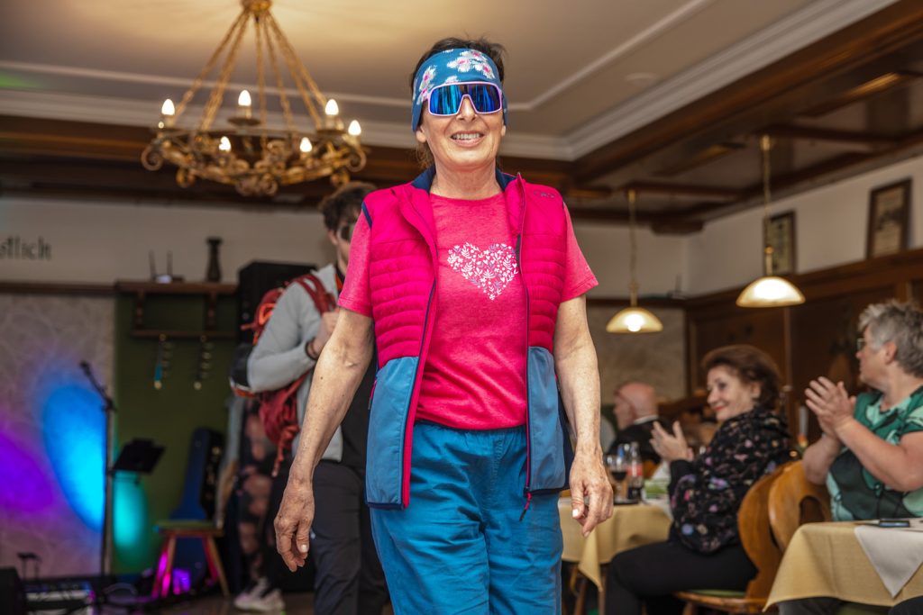 Eine Frau präsentiert farbenfrohe Outdoor- und Freizeitmode von Sport Spanner bei einer Modenschau. Sie trägt ein pinkes Sportshirt, eine passende Steppweste, blaue Outdoorhose sowie ein Stirnband und eine große Sportsonnenbrille. Die Gäste sitzen entlang des Laufstegs im gemütlichen Gastraum und applaudieren der Präsentation.