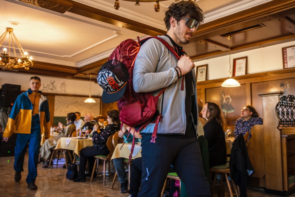 Ein junger Mann präsentiert funktionelle Outdoor- und Wanderbekleidung von Sport Spanner bei einer Modenschau. Er trägt eine sportliche Jacke, dunkle Outdoorhose, große Sportsonnenbrille und einen großen Wanderrucksack mit Cap. Im Hintergrund läuft ein weiteres Model mit farbenfroher Outdoorbekleidung durch den Gastraum, während die Gäste die Show verfolgen.