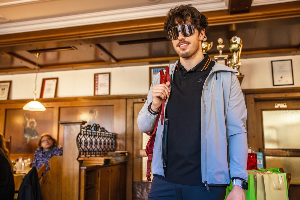 Ein junger Mann präsentiert moderne Outdoor- und Freizeitmode von Sport Spanner bei einer Modenschau. Er trägt eine sportliche Jacke, ein dunkles Poloshirt, eine große Sportsonnenbrille und einen Rucksack über der Schulter. Die Gäste beobachten die Präsentation im stilvollen Gastraum aufmerksam.