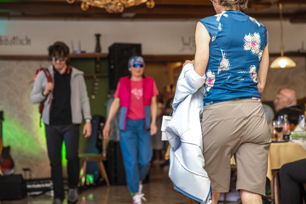 Mehrere Personen präsentieren funktionelle Outdoor- und Sportbekleidung von Sport Spanner bei einer Modenschau. Im Vordergrund ist ein sportliches Outfit mit ärmellosem Funktionsshirt und Shorts zu sehen, während weitere Models mit Wander- und Freizeitmode den Laufsteg entlanggehen. Die Gäste verfolgen die Show aufmerksam im gemütlichen Gastraum.
