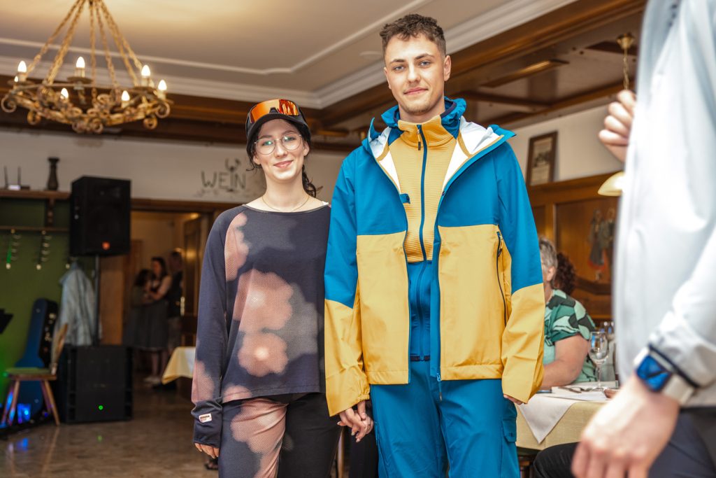 Ein junges Paar präsentiert moderne Outdoor- und Wintersportmode von Sport Spanner bei einer Modenschau. Die Frau trägt ein lässiges Sport-Outfit mit Cap und Brille, der Mann eine farbenfrohe Ski- bzw. Outdoorjacke mit passender Hose. Beide stehen Hand in Hand im Mittelpunkt des Gastraums, während die Gäste die Präsentation verfolgen.