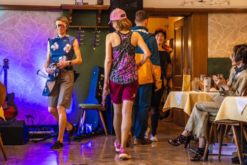 Mehrere Personen präsentieren sportliche Outdoor- und Laufbekleidung bei einer Modenschau von Sport Spanner. Zu sehen sind funktionelle Shirts, Shorts, Jacken, Caps und Sportbrillen in kräftigen Farben. Die Models gehen durch den Gastraum, während die Gäste am Rand applaudieren und die Sportmode aufmerksam betrachten.