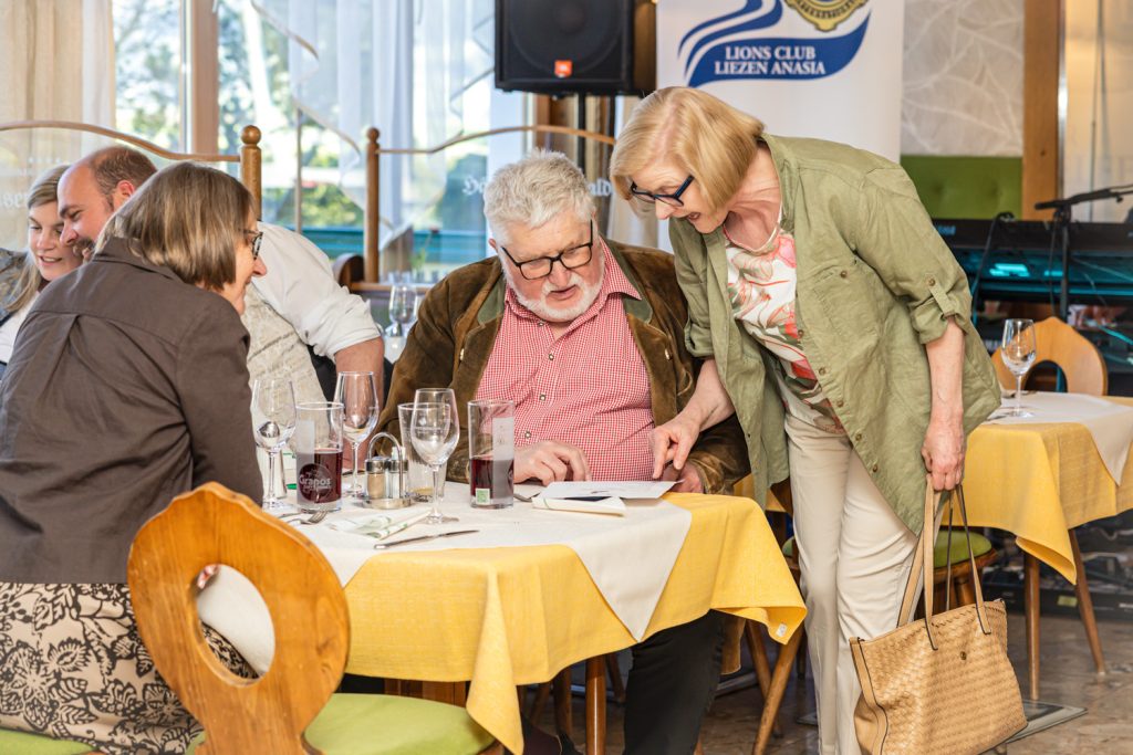 Mehrere Gäste sitzen gemeinsam an einem gedeckten Tisch in einem Restaurant und schauen auf ein Blatt Papier. Eine Frau im grünen Hemd beugt sich interessiert vor und zeigt auf das Dokument, während ein älterer Herr aufmerksam mitliest. Die Szene wirkt freundlich, aufmerksam und gesellig.