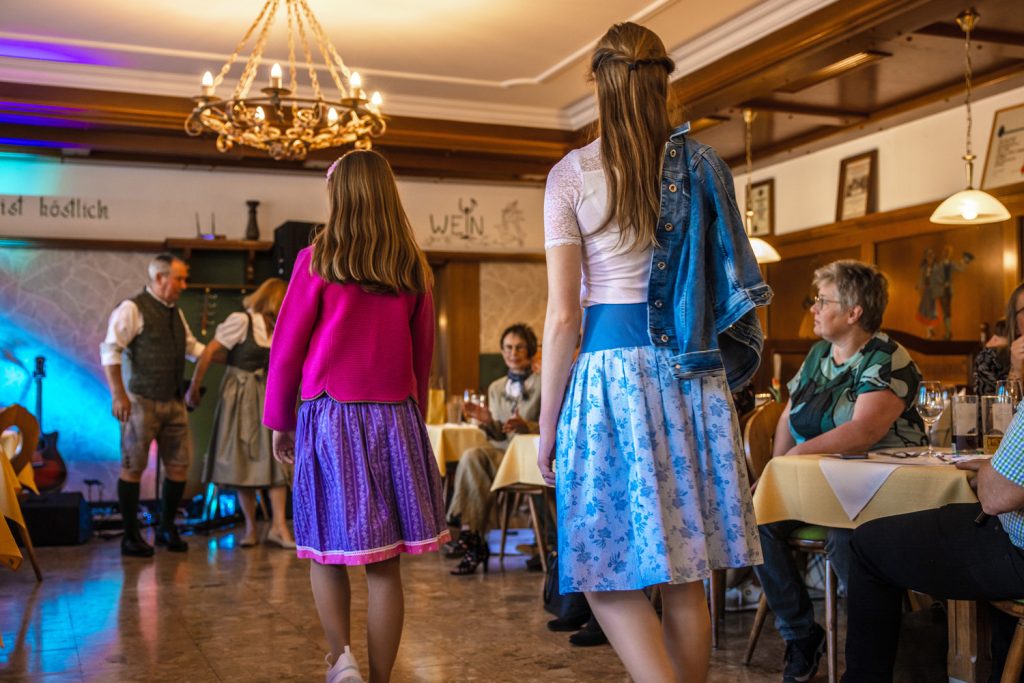 Zwei Mädchen präsentieren Trachtenmode bei einer Modenschau in einem gemütlichen Gasthaus. Das ältere Mädchen trägt ein blau-weiß gemustertes Kleid mit Jeansjacke über der Schulter, das jüngere ein lila Kleid mit pinker Strickjacke. Die Gäste sitzen entlang des Laufstegs und verfolgen die Präsentation aufmerksam.
