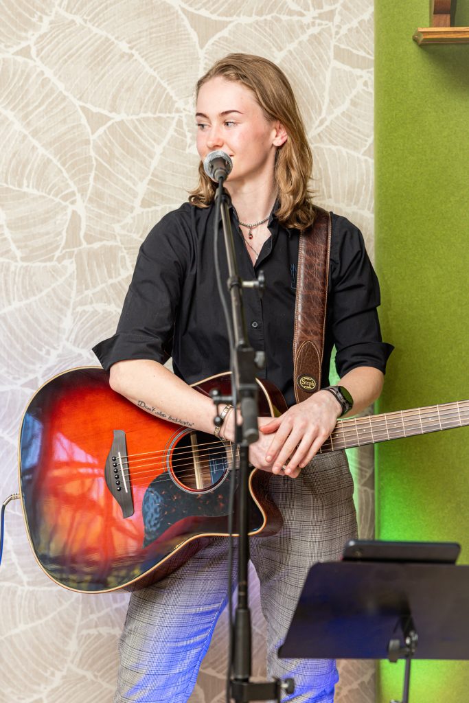 Eine junge Musikerin steht mit einer akustischen Gitarre vor einem Mikrofon und lächelt leicht zur Seite. Sie trägt ein schwarzes Hemd und karierte Hosen, während die Bühne dezent mit grünem Licht beleuchtet wird. Die Atmosphäre wirkt entspannt und persönlich.