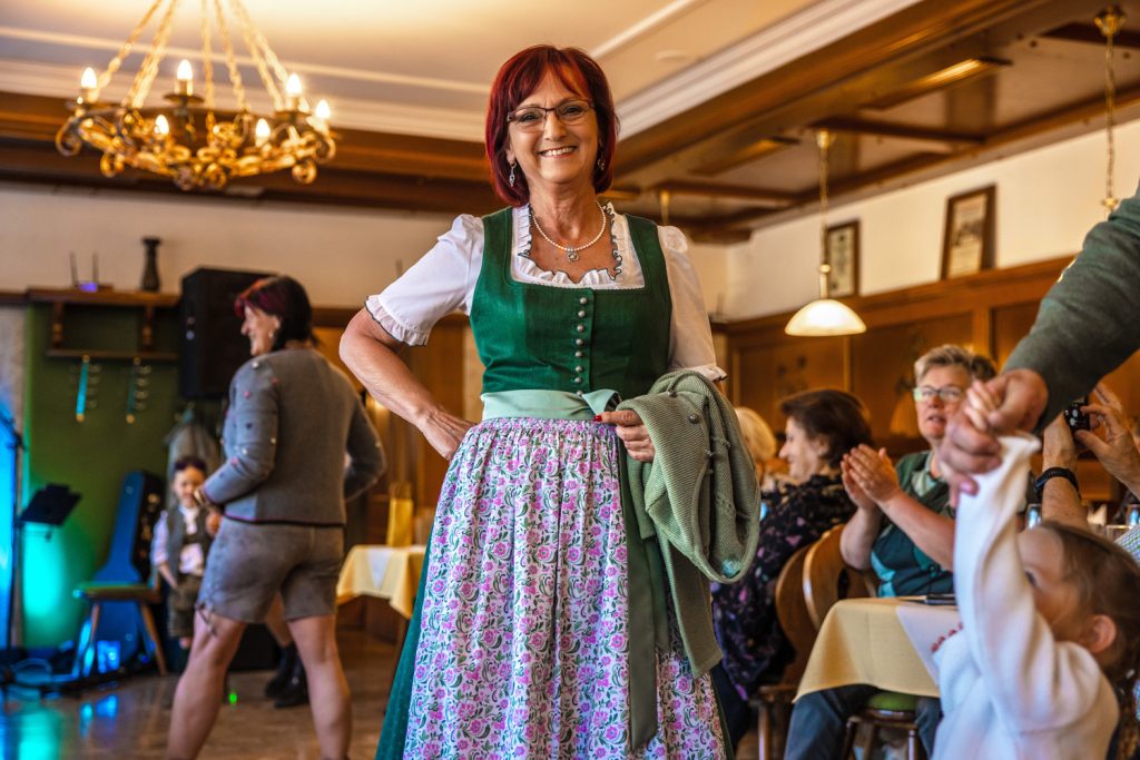 Eine Frau präsentiert ein traditionelles grünes Dirndl mit floral gemustertem Rock bei einer Modenschau in einem gemütlichen Gasthaus. Sie hält eine passende grüne Strickjacke in der Hand und lächelt in die Kamera, während die Gäste entlang des Laufstegs applaudieren und zuschauen.