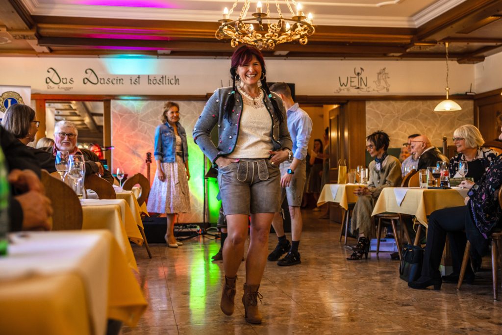 Eine Frau präsentiert moderne Trachtenmode auf dem Laufsteg eines gemütlichen Gasthauses. Sie trägt eine graue kurze Lederhose, eine verzierte graue Strickjacke, ein weißes Spitzentop und braune Stiefeletten. Im Hintergrund warten weitere Teilnehmer der Modenschau, während die Gäste an den Tischen die Präsentation aufmerksam verfolgen.