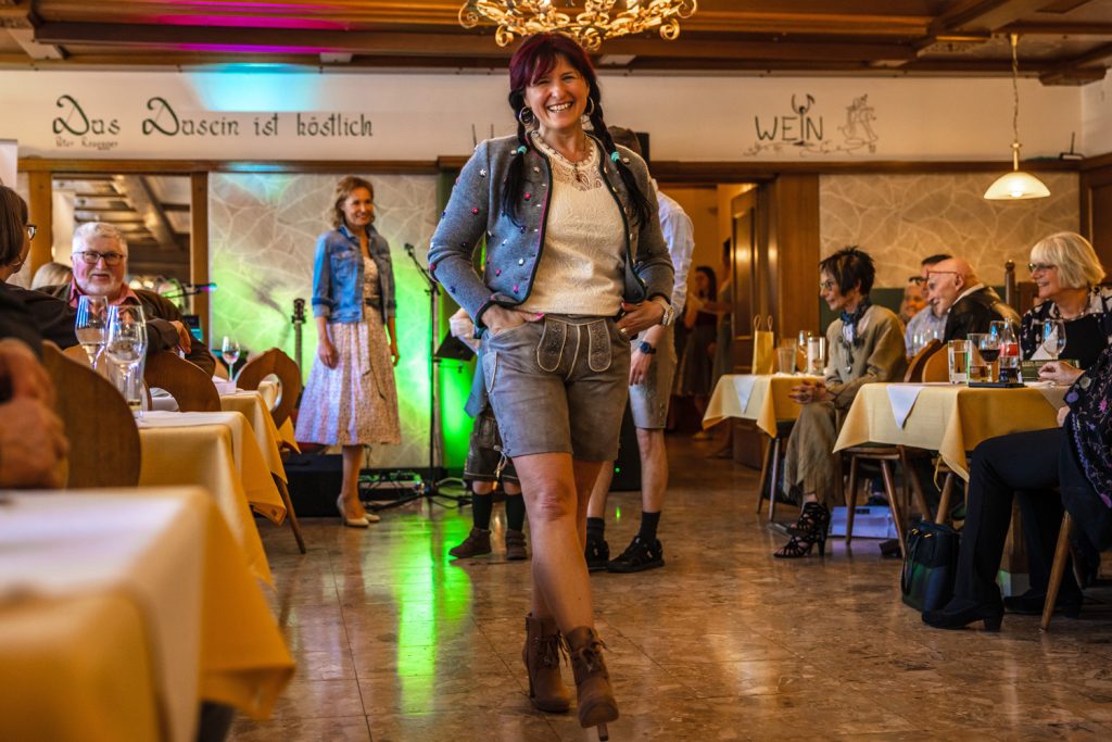 Eine Frau präsentiert moderne Trachtenmode bei einer Modenschau in einem traditionellen Gasthaus. Sie trägt eine graue kurze Lederhose, eine verzierte Strickjacke, ein weißes Spitzentop und braune Stiefeletten. Im Hintergrund stehen weitere Models bereit, während die Gäste entlang des Laufstegs die Show aufmerksam verfolgen.