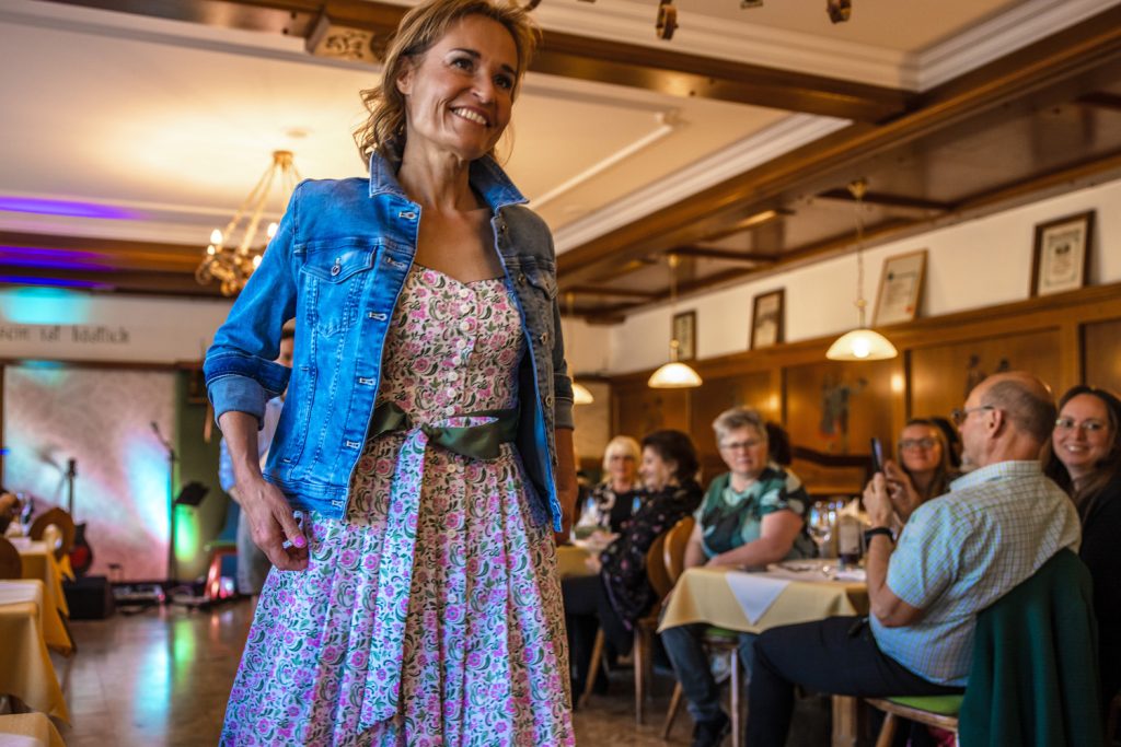 Eine Frau präsentiert bei einer Modenschau ein florales Dirndl mit grünem Gürtel und Jeansjacke. Sie läuft lächelnd durch ein gemütliches Gasthaus, während die Gäste an den Tischen sitzen, zuschauen und Fotos machen.