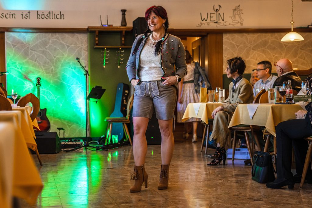 Eine Frau präsentiert moderne Trachtenmode bei einer Modenschau in einem gemütlichen Gasthaus. Sie trägt eine graue kurze Lederhose, eine verzierte graue Jacke, ein weißes Spitzentop und braune Stiefeletten. Die Gäste sitzen an den Tischen entlang des Laufstegs und beobachten die Präsentation aufmerksam.