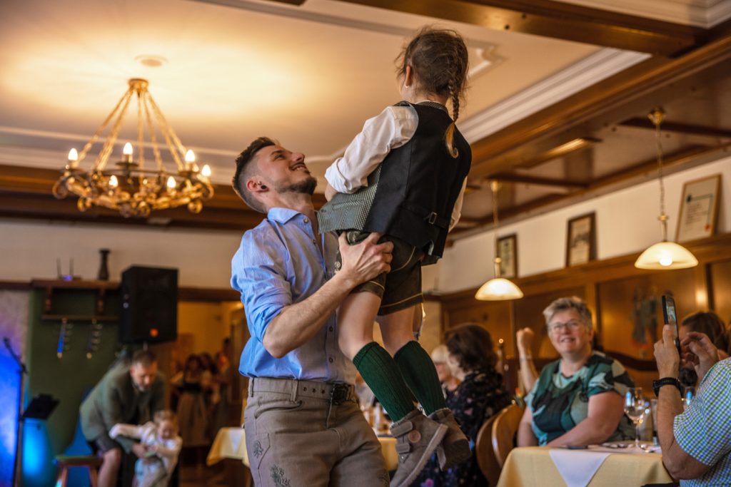 Ein Mann hebt ein kleines Mädchen in traditioneller Trachtenkleidung bei einer Modenschau in einem gemütlichen Gasthaus hoch. Das Mädchen trägt Lederhose, grüne Kniestrümpfe und eine Weste, während die Gäste rundherum lächeln, applaudieren und den besonderen Moment genießen.