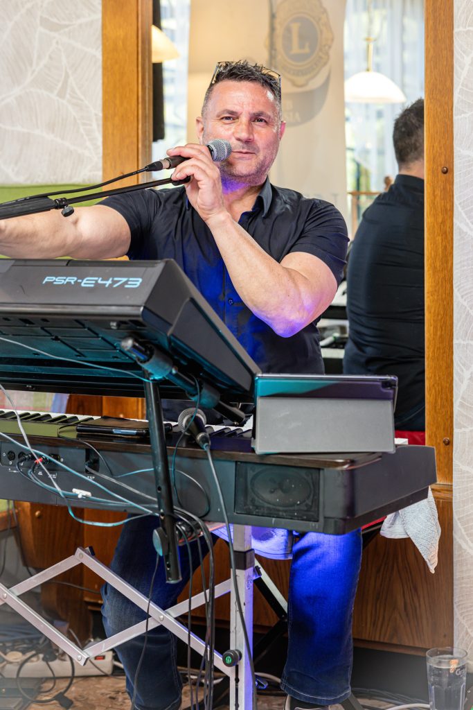 Ein Musiker sitzt hinter einem Keyboard und spricht oder singt in ein Mikrofon. Er trägt ein dunkles Hemd und blickt direkt in die Kamera. Vor ihm stehen mehrere technische Geräte und Kabel, während im Hintergrund Spiegel und Holzelemente des Veranstaltungsraums zu sehen sind.