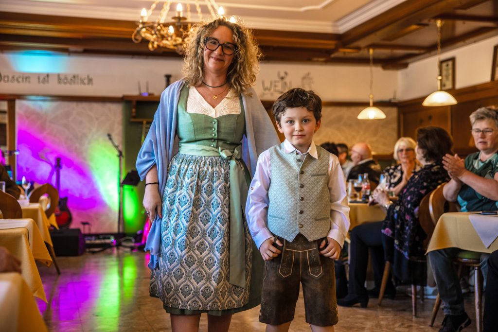 Eine Frau und ein kleiner Junge stehen gemeinsam auf dem Laufsteg einer Trachtenmodenschau in einem gemütlichen Restaurant. Die Frau trägt ein grün-blau gemustertes Dirndl mit hellblauem Schultertuch, der Junge eine Lederhose mit hellgrüner Weste und weißem Hemd. Im Hintergrund sitzen Gäste an festlich gedeckten Tischen und applaudieren den beiden.