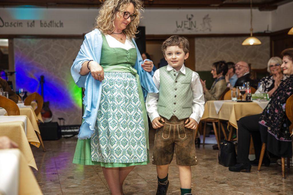 Eine Frau und ein kleiner Junge präsentieren traditionelle Trachtenmode bei einer Modenschau in einem gemütlichen Gasthaus. Die Frau trägt ein grün gemustertes Dirndl mit hellblauem Schultertuch, der Junge eine Lederhose mit hellgrüner Weste. Beide gehen gemeinsam über den Laufsteg, während die Gäste an den Tischen lächeln und applaudieren.