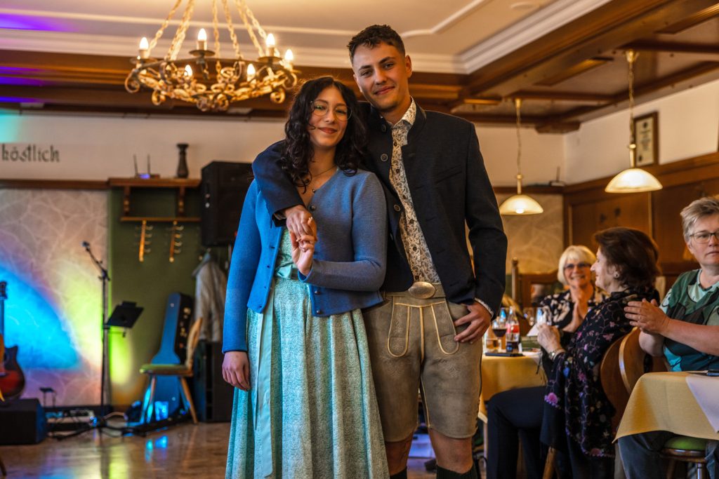 Ein junges Paar präsentiert Trachtenmode bei einer Modenschau in einem gemütlichen Gasthaus. Die Frau trägt ein mintgrünes Kleid mit blauer Strickjacke und der Mann Lederhose mit dunklem Trachtenjacket. Beide stehen eng beieinander und lächeln entspannt in die Kamera, während die Gäste an den Tischen die Modenschau aufmerksam verfolgen und applaudieren.