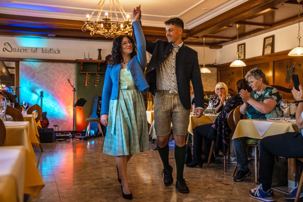Ein junges Paar präsentiert Trachtenmode bei einer Modenschau in einem gemütlichen Gasthaus. Die Frau trägt ein mintgrünes Kleid mit blauer Strickjacke und schwarze Pumps, während der Mann Lederhose, dunkles Trachtenjacket und Kniestrümpfe trägt. Gemeinsam gehen sie lächelnd über den Laufsteg, halten die Hände erhoben und werden von den Gästen mit Applaus begleitet.