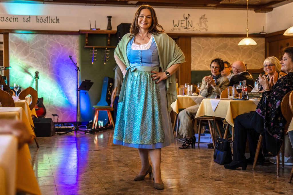 Eine Frau präsentiert ein elegantes Dirndl bei einer Modenschau in einem gemütlichen Gasthaus. Sie trägt ein blau-grünes Dirndl mit gemusterter Schürze, einen hellgrünen Schal über den Schultern und Pumps. Mit den Händen in der Hüfte steht sie selbstbewusst auf dem Laufsteg, während die Gäste an den Tischen applaudieren und die Trachtenmode aufmerksam betrachten.
