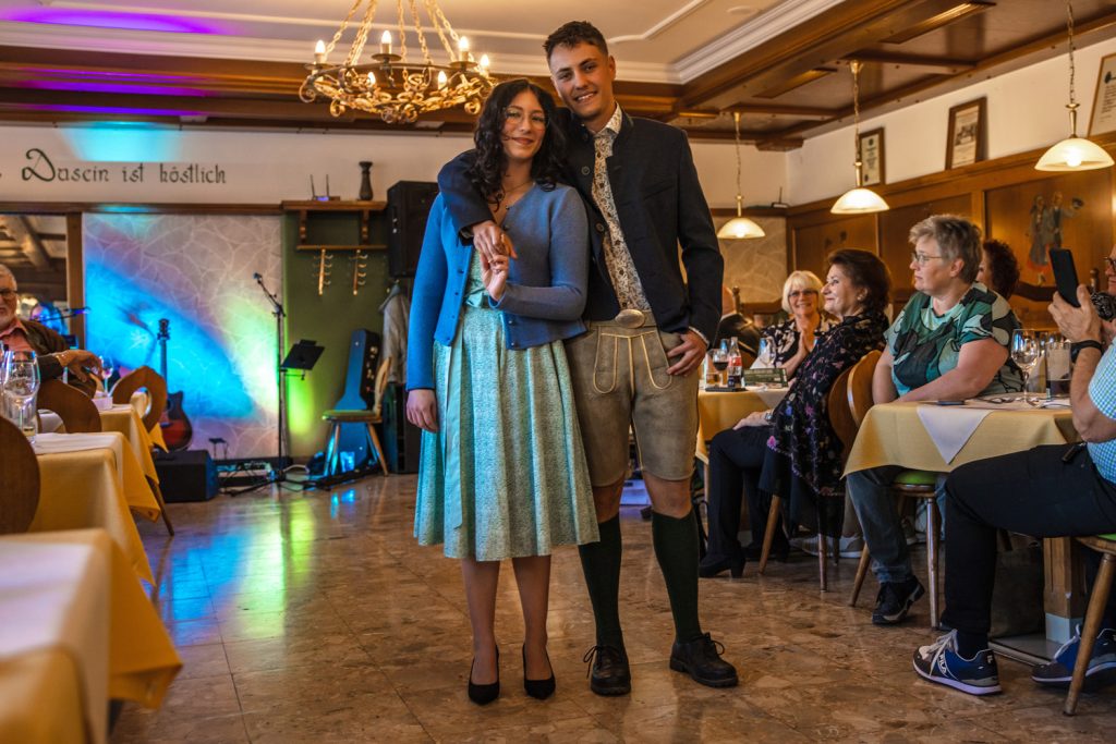 Ein junges Paar präsentiert Trachtenmode bei einer Modenschau in einem gemütlichen Gasthaus. Die Frau trägt ein mintgrünes Kleid mit blauer Strickjacke und schwarzen Pumps, während der Mann Lederhose, Kniestrümpfe und ein dunkles Trachtenjacket trägt. Beide stehen eng beieinander und lächeln in die Kamera, während die Gäste an den Tischen die Modenschau aufmerksam verfolgen.