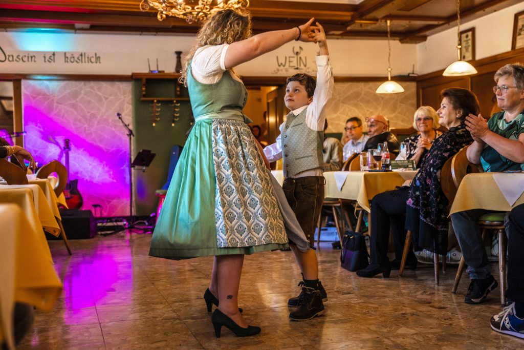 Eine Frau und ein Junge in traditioneller Trachtenmode tanzen gemeinsam bei einer Modenschau in einem gemütlichen Gasthaus. Die Frau trägt ein grünes Dirndl mit gemustertem Rock und schwarze Pumps, während der Junge Lederhose, Hemd und Weste trägt und ihre Hand zum Drehen hält. Die Gäste sitzen entlang des Laufstegs, applaudieren und beobachten die Szene aufmerksam, während bunte Lichteffekte und die rustikale Einrichtung eine festliche Atmosphäre schaffen.
