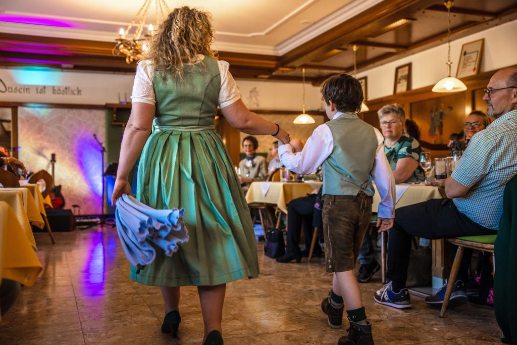 Eine Frau und ein Junge in traditioneller Trachtenkleidung gehen Hand in Hand über den Laufsteg einer Modenschau in einem gemütlichen Gasthaus. Die Frau trägt ein grünes Dirndl und hält ein hellblaues Tuch in der Hand, während der Junge Lederhose, Hemd und Weste trägt. Die Gäste sitzen an den Tischen entlang des Raumes und verfolgen die Szene aufmerksam, während bunte Lichteffekte und die rustikale Holzeinrichtung für eine festliche Stimmung sorgen.